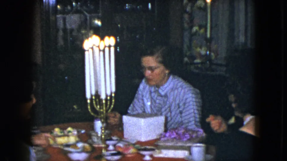 1951 Cleveland dining-room frame with a woman in a striped blouse, tall candelabra, food plates, cups, flowers, and nearby guests.