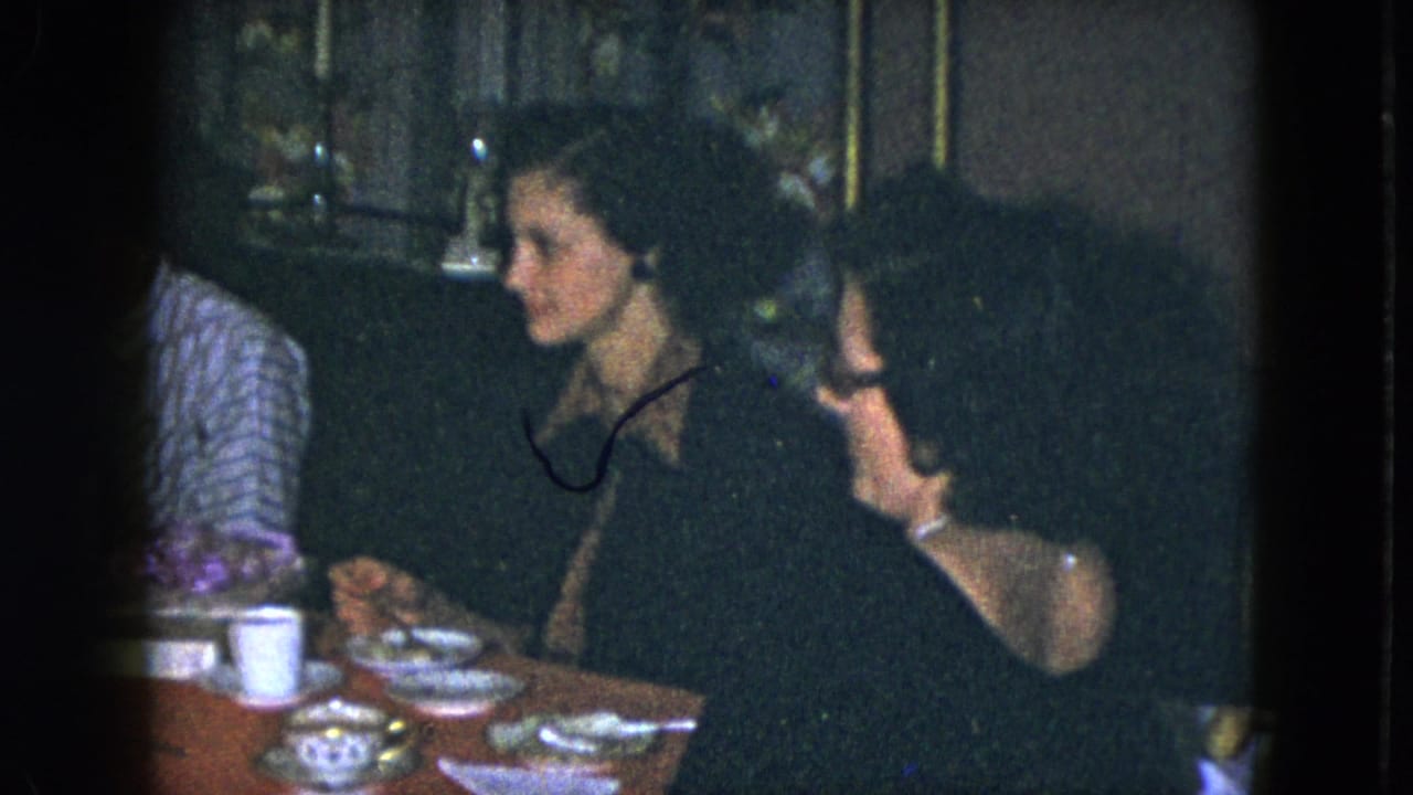Women converse at a table set with cups, saucers, plates, and small dishes in a dark room.
