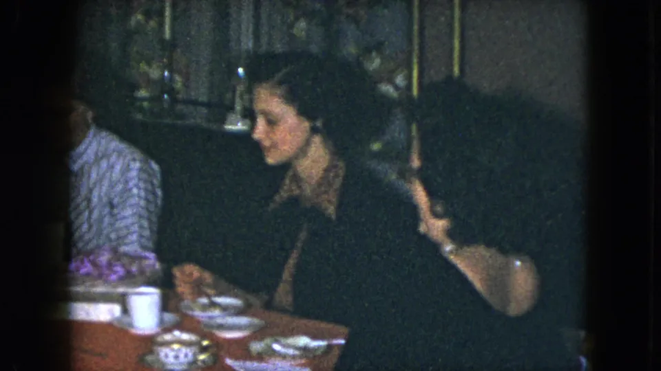 1951 Cleveland home-movie frame showing seated women at a red-covered dining table with cups, plates, and dim interior furnishings.