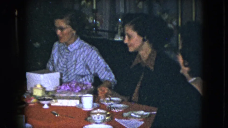1951 Cleveland home-movie frame with an elderly woman and another woman seated beside a red tablecloth, teacups, plates, and a white gift box.
