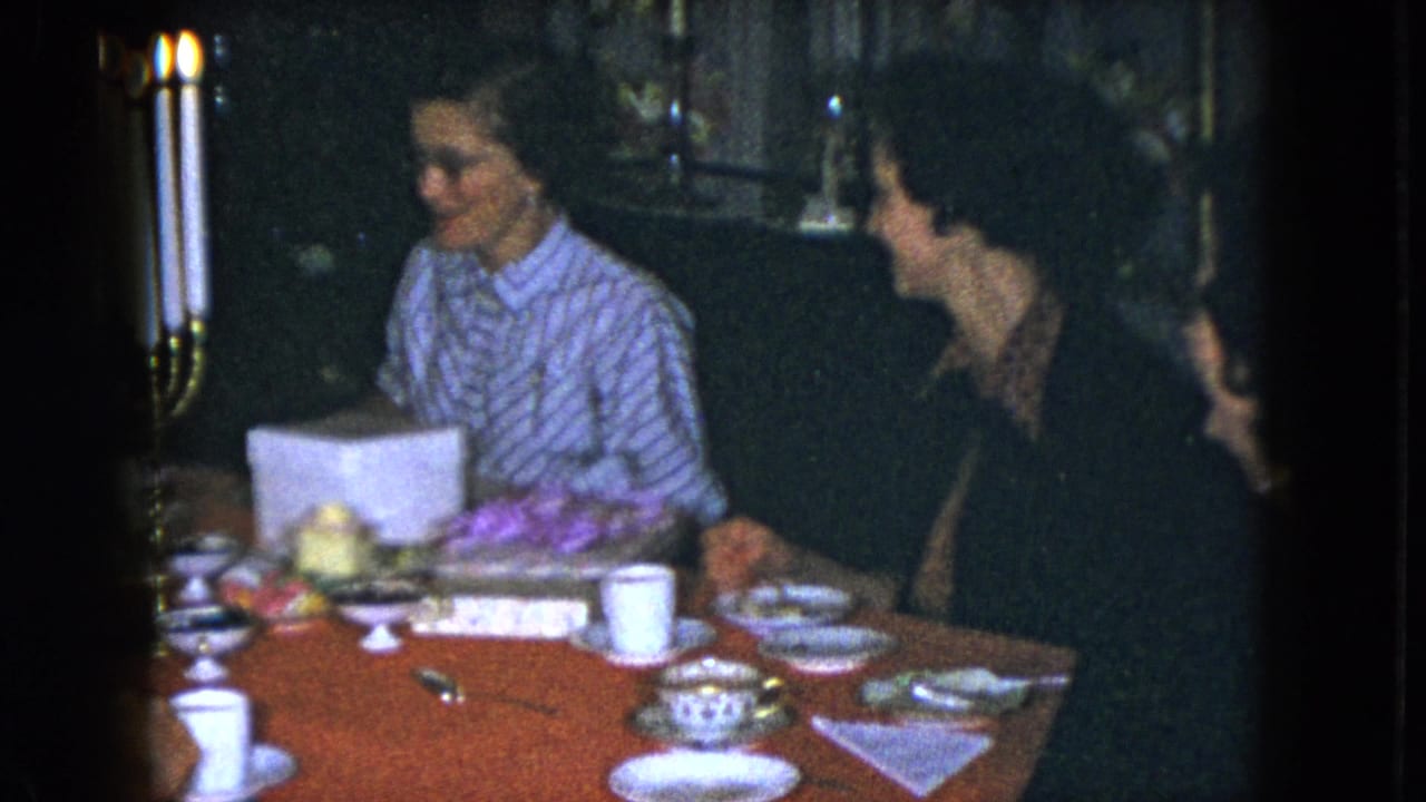 In this Cleveland 1951 home-movie frame, adult women sit at a red-covered table while one woman handles a white gift box near a tall candelabra.