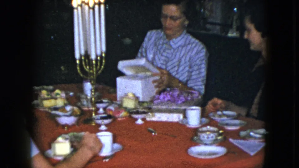 During the Cleveland 1951 home birthday scene, a woman lifts wrapping from a white gift box while other adults sit nearby at a candlelit table.