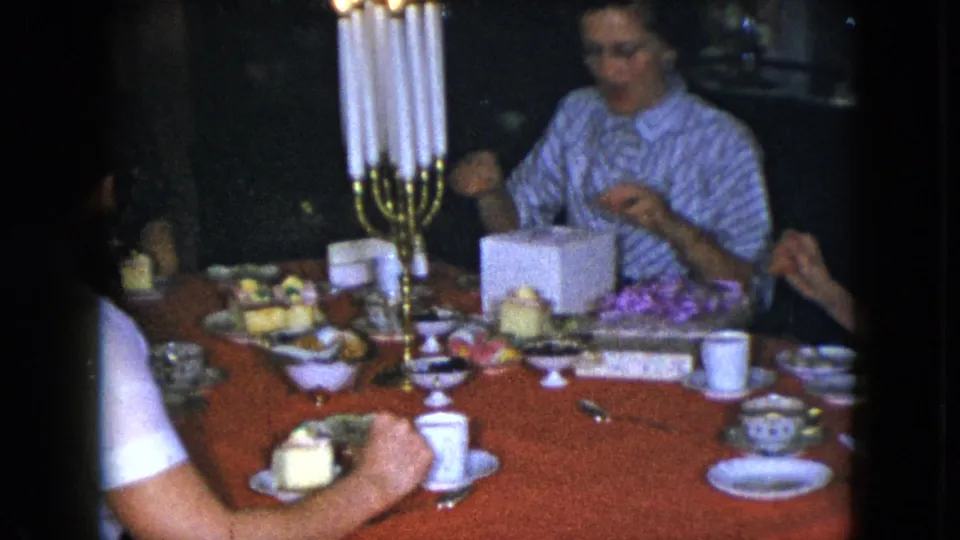 The Cleveland 1951 home-movie frame shows adult women around a red tablecloth with a tall candelabra, desserts, cups, and an open gift box.