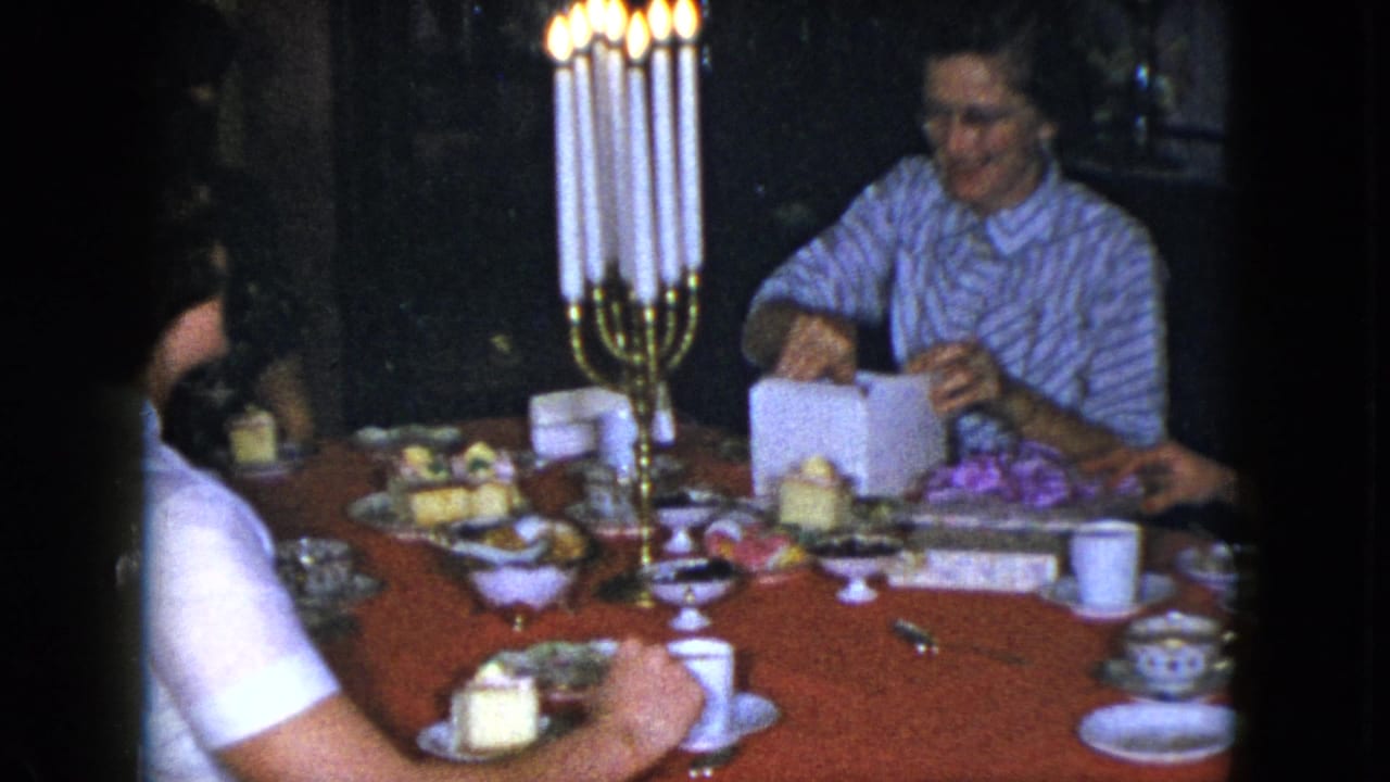 1951 Cleveland home-movie frame of adults at a red-covered dinner table with lit candelabra as a woman handles a box.