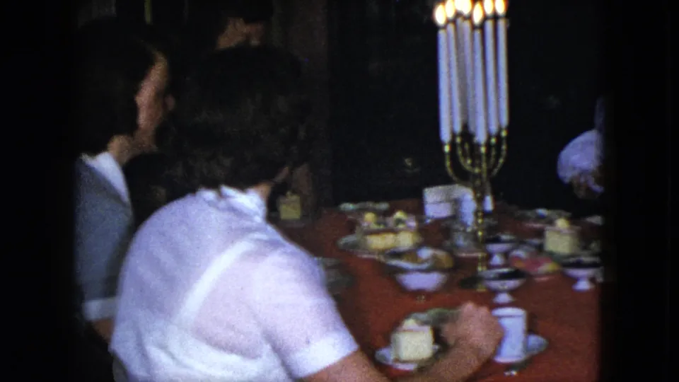 Cleveland 1951 dining-room frame showing adults around a red tablecloth, lit candelabra, cups, and dessert plates.
