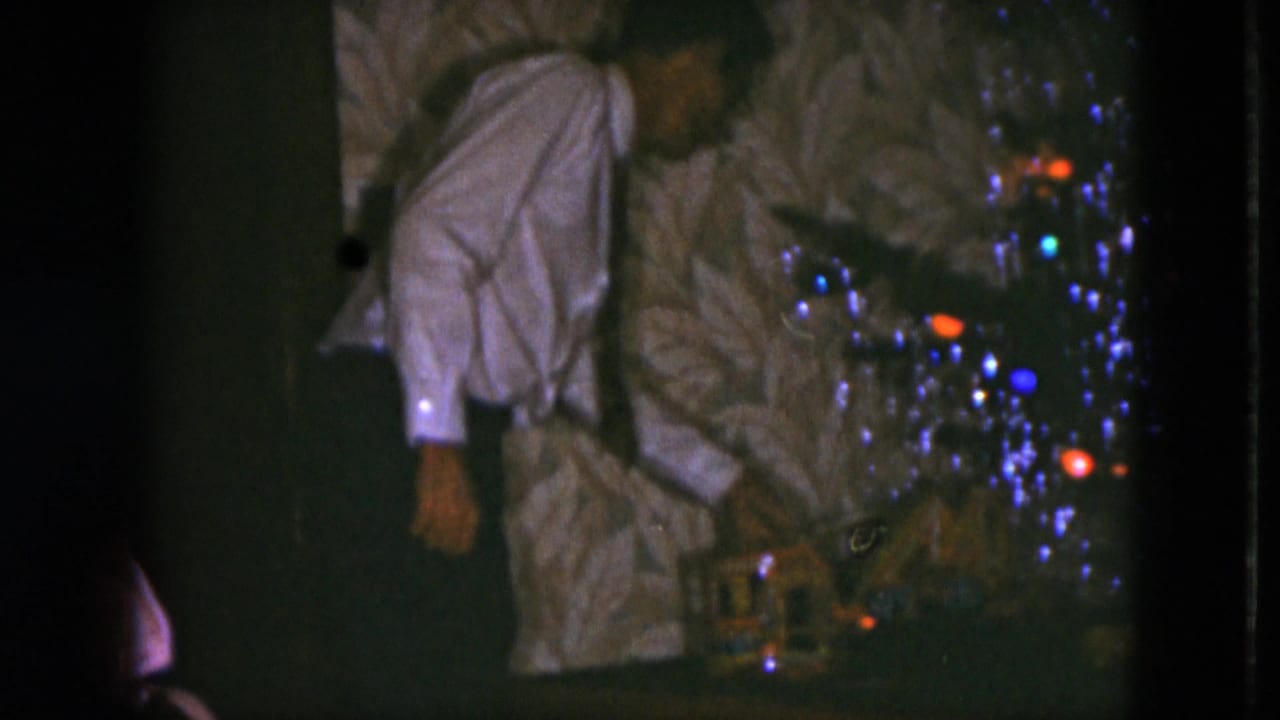 Man in a white shirt leans over a model train beneath a decorated Christmas tree.