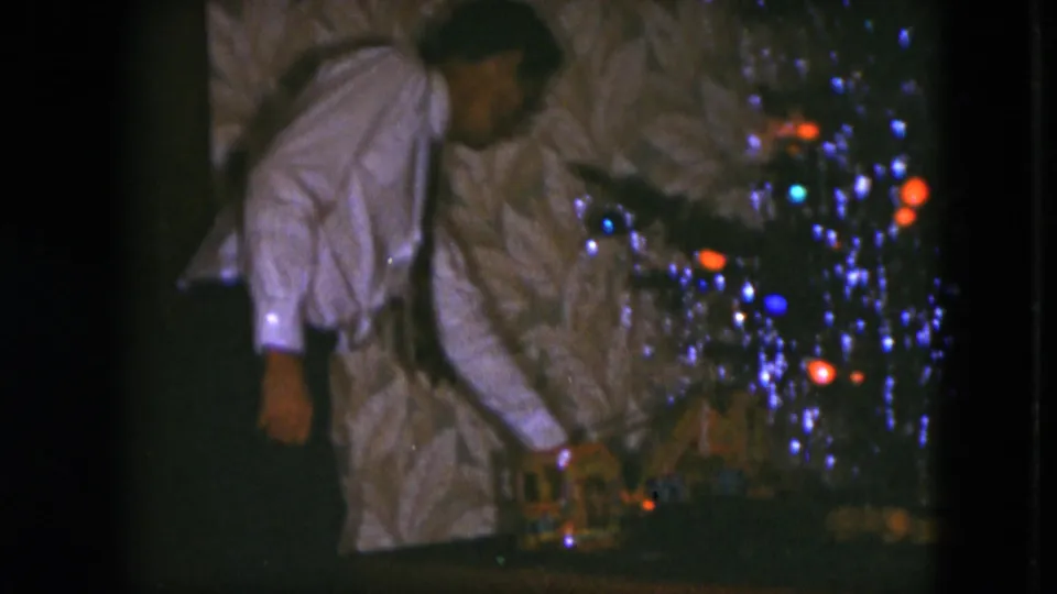 The man remains bent toward the small train set beneath the decorated tree, with colored lights and reflective tinsel visible in the Cleveland interior.
