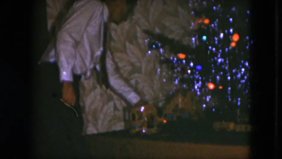 A low-light 8mm frame shows the man leaning beside the tinsel-covered tree, with the model train and track partly visible below.