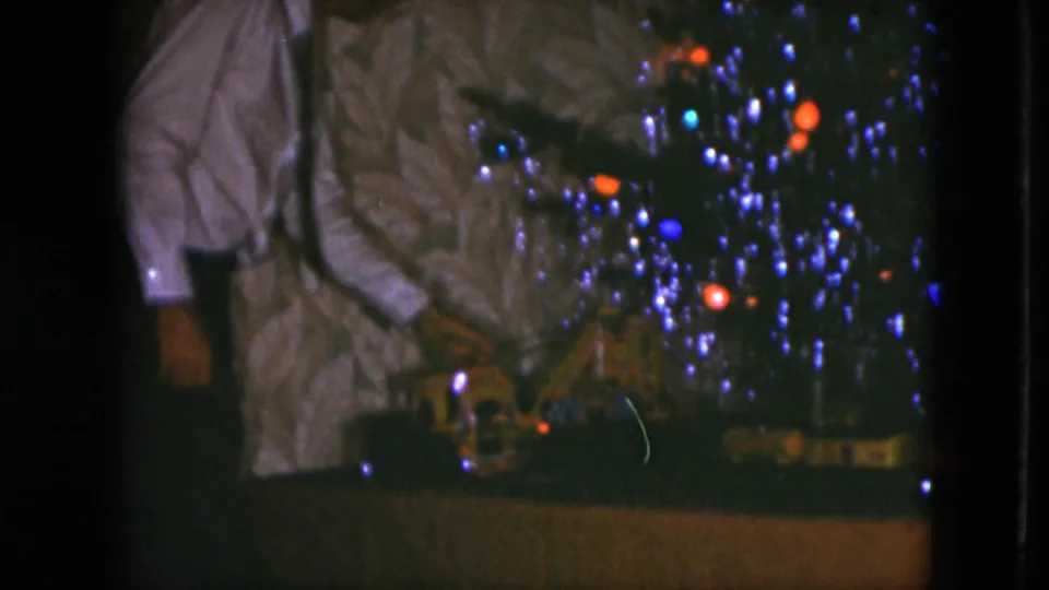 The final frame shows the man reaching near the model train under the Christmas tree, with bright colored bulbs and tinsel against patterned wallpaper.