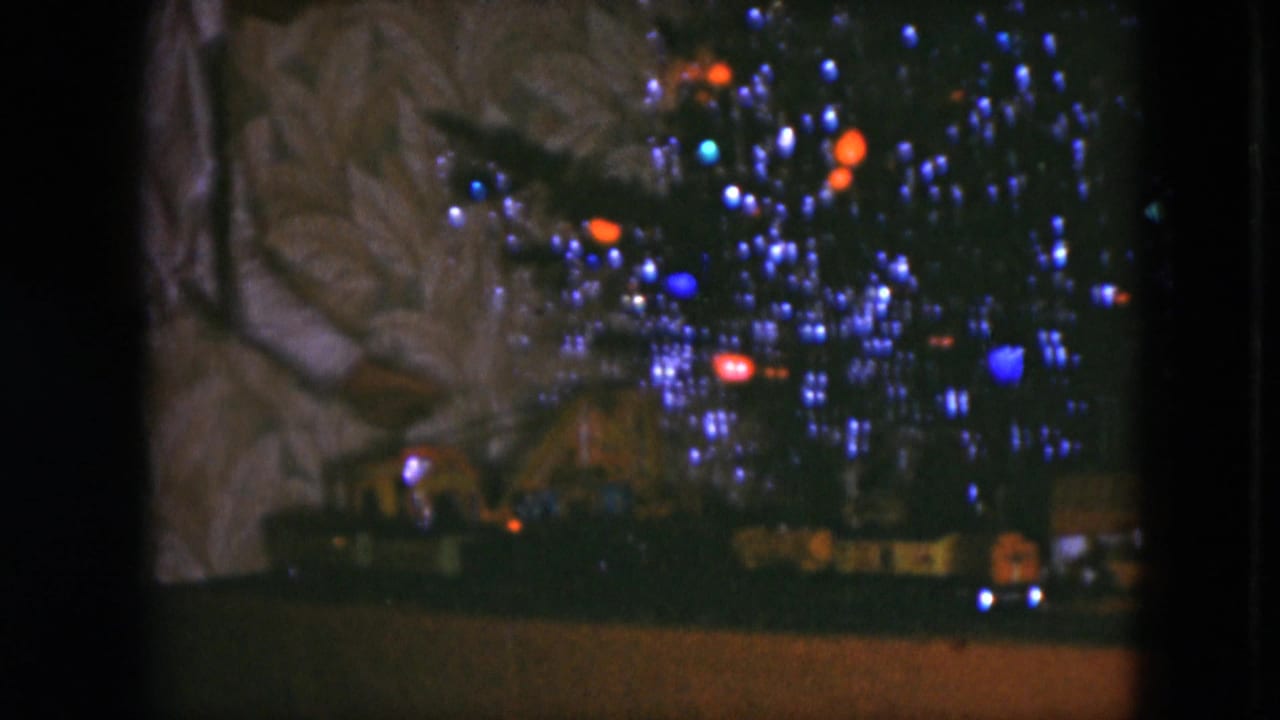 Lit Christmas tree with colored bulbs above a toy train layout in a dark interior.