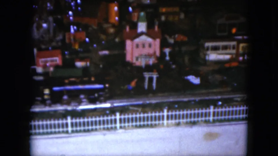 1951 Cleveland home-movie frame showing a dark indoor toy train village with a pink church-like building and white picket fence.