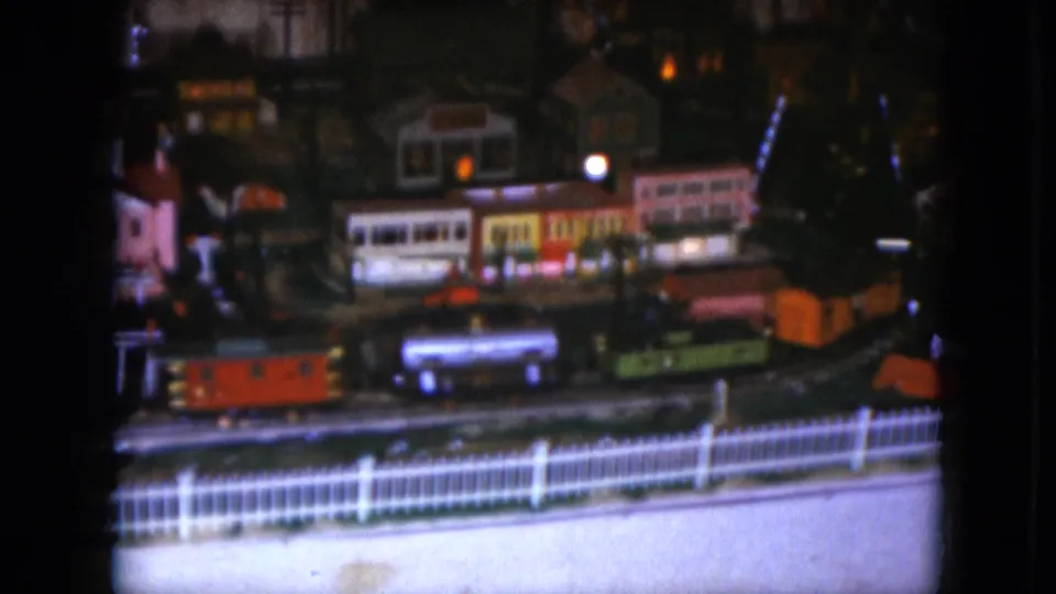 1951 Cleveland home-movie frame showing orange, blue, and green toy train cars passing a row of miniature village buildings.