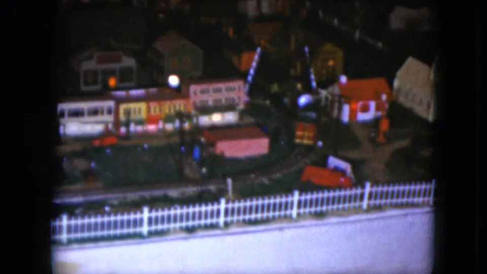 1951 Cleveland frame showing a toy train turning through a miniature village with small buildings, lights, and a foreground white fence.