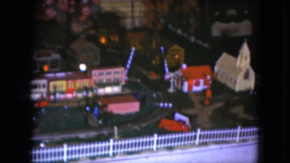 Home-movie view of a Cleveland model train layout, showing illuminated miniature buildings, a church, and a fence along the foreground.