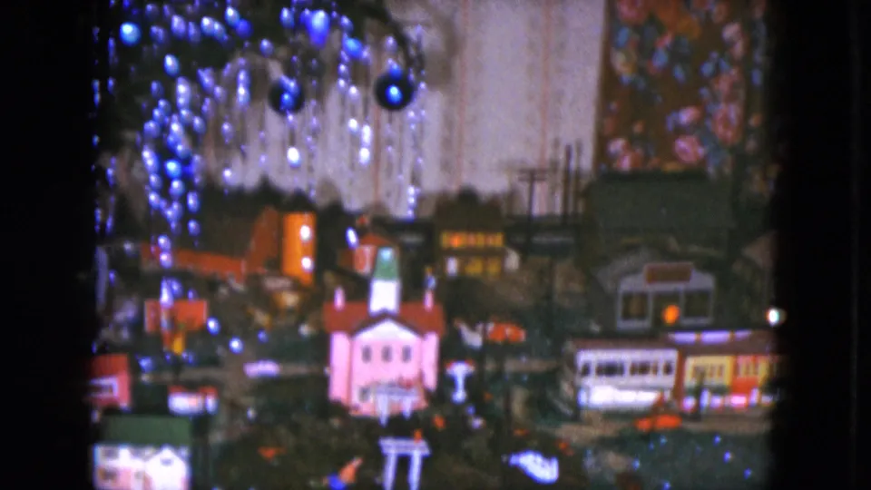 1951 Cleveland 8mm frame showing the holiday model railroad from a shifted angle, with lit buildings and blue ornaments above the miniature town.