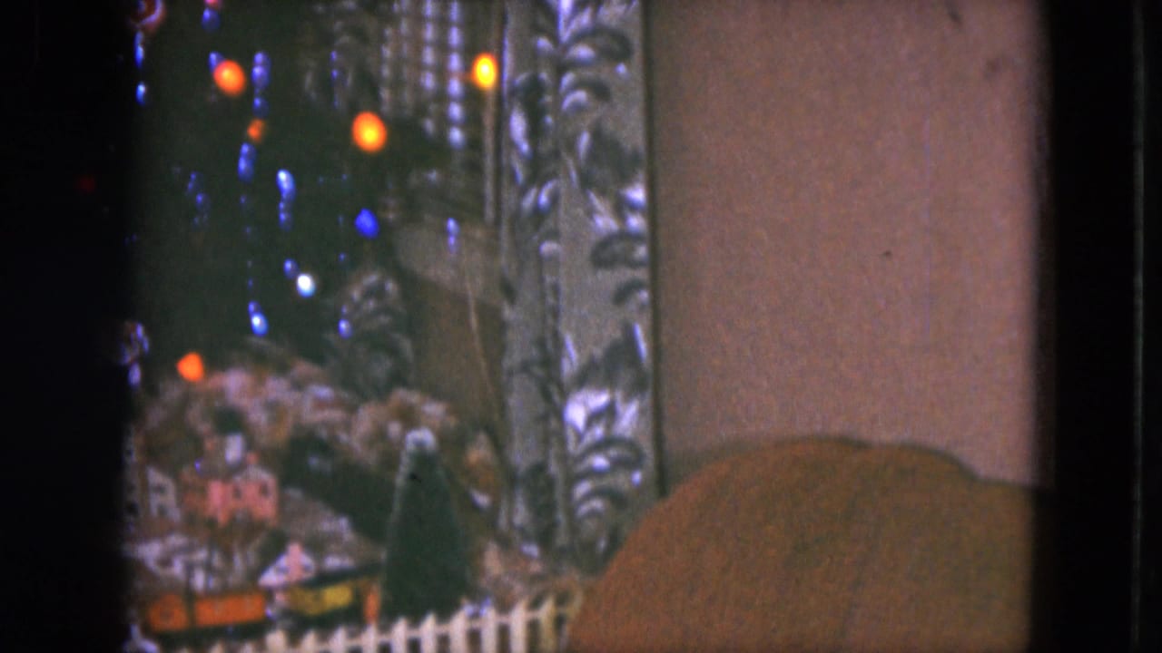 1951 Cleveland home-movie frame showing miniature train scenery under a Christmas tree with colored lights and patterned curtains.