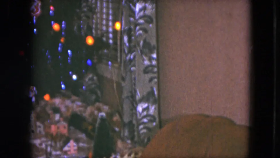 1951 Cleveland home-movie frame with glowing tree lights, small train-set scenery, patterned curtains, and a plain wall area.