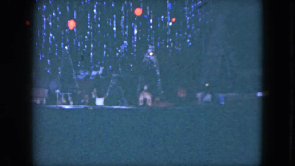 Home-movie frame showing the base of a Christmas tree with ornaments, miniature decorations, and the train layout below.