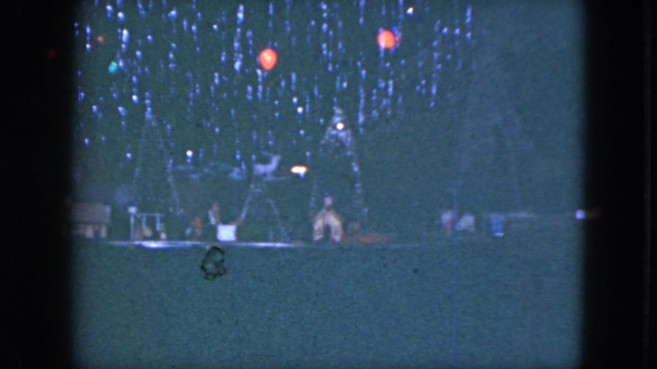 1951 Cleveland home-movie frame showing a dim holiday model train display with trees, small buildings, and red lights.