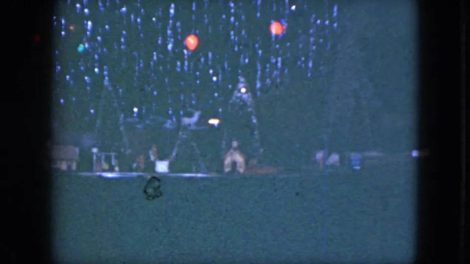 1951 Cleveland home-movie frame showing a dim holiday model train display with trees, small buildings, and red lights.