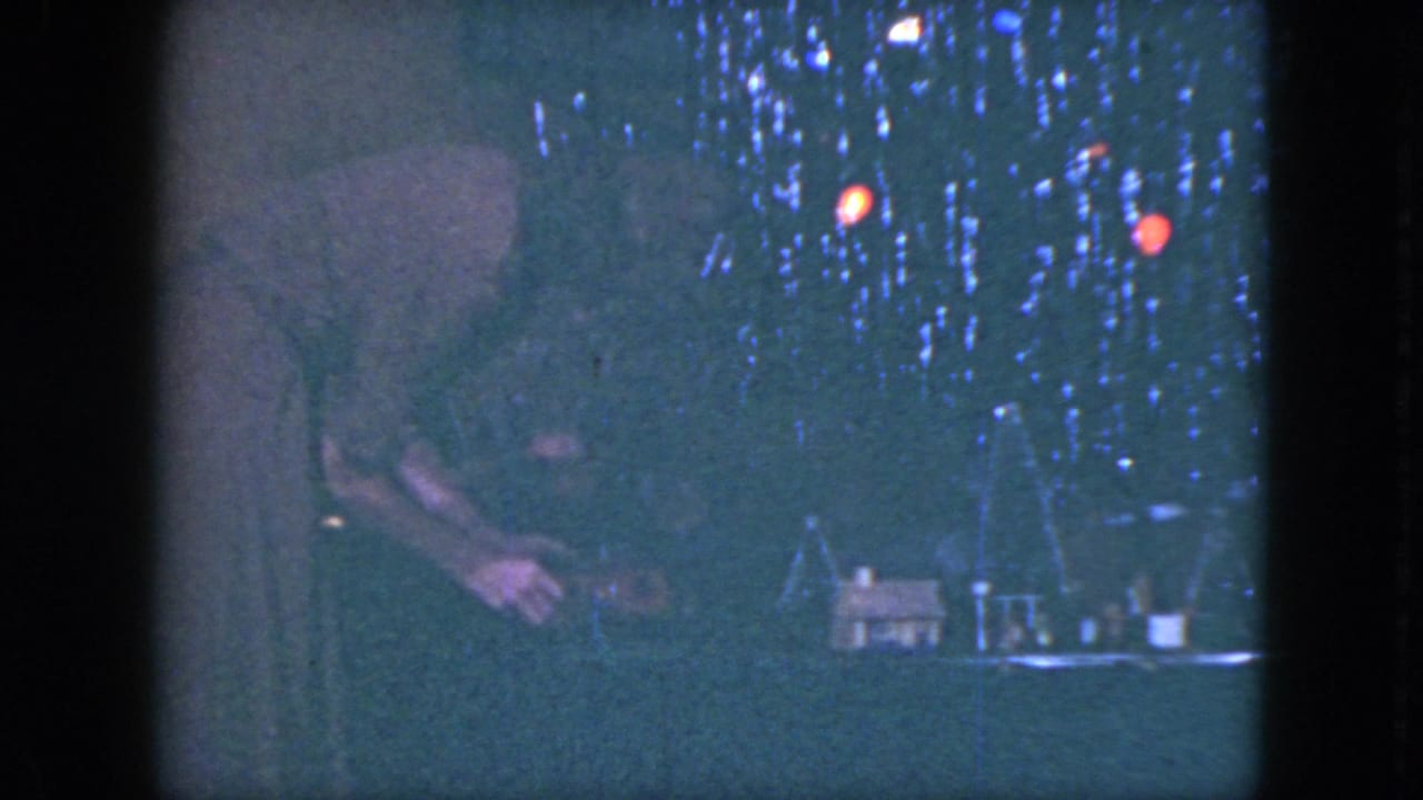 In 1951 Cleveland home-movie footage, a woman leans into a dim model train display with miniature houses and glowing holiday lights.