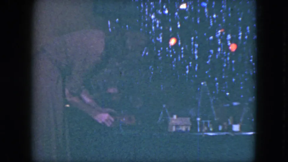 In 1951 Cleveland home-movie footage, a woman leans into a dim model train display with miniature houses and glowing holiday lights.