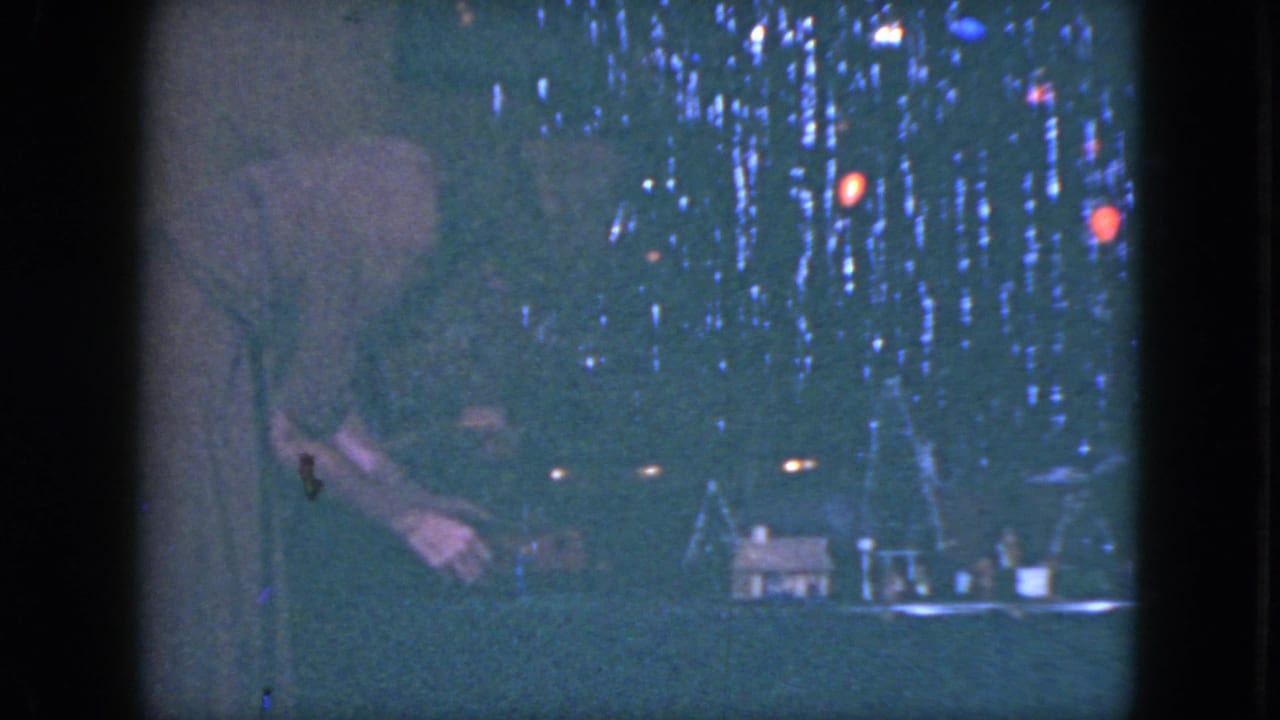 A woman stands at the left of a Cleveland 1951 model train village, reaching near small houses under red and blue holiday lights.