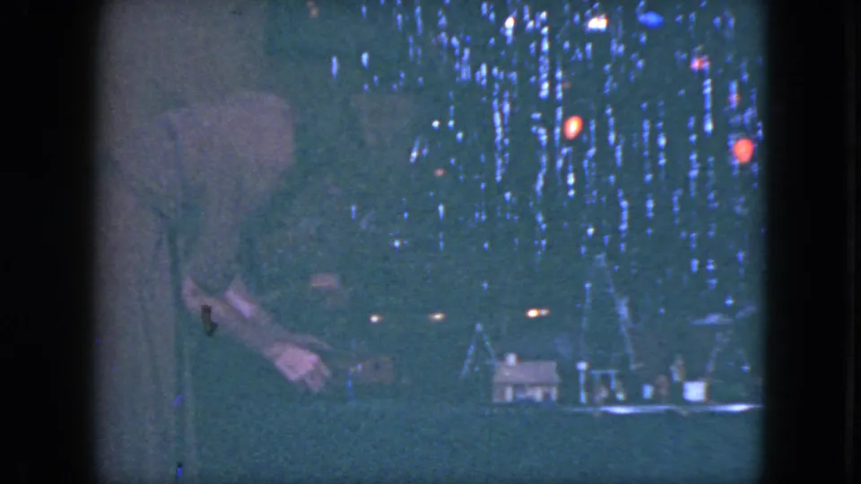 A woman stands at the left of a Cleveland 1951 model train village, reaching near small houses under red and blue holiday lights.