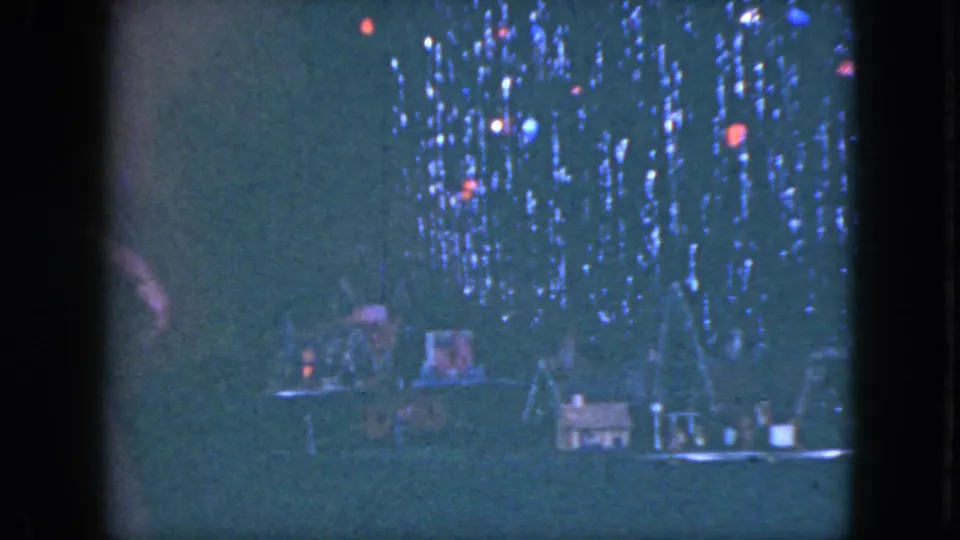 The model train-set village is seen in dim 8mm footage, with small structures and glowing colored lights as a hand moves out of frame.