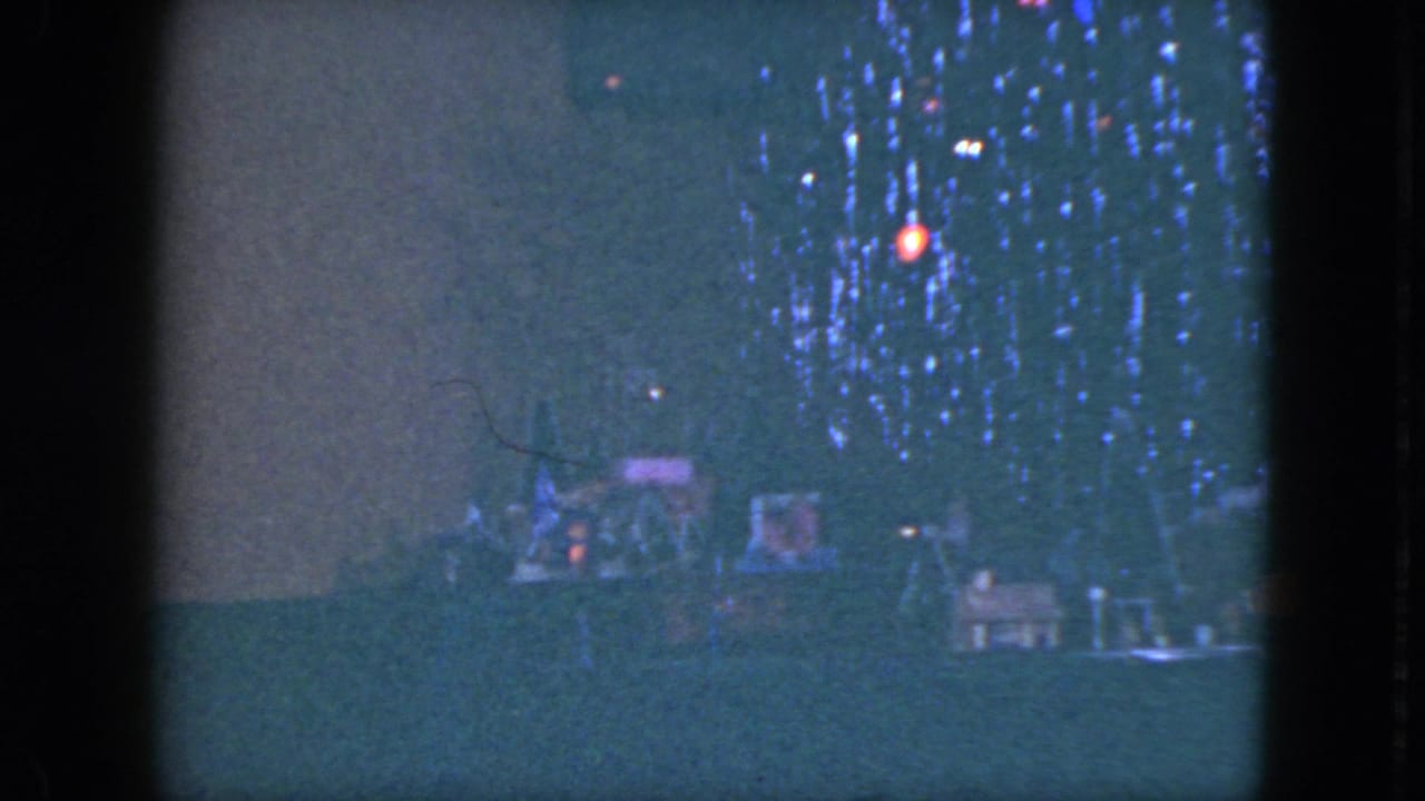 1951 Cleveland home-movie frame showing a dim Christmas tree with lights and a tabletop train set nearby.