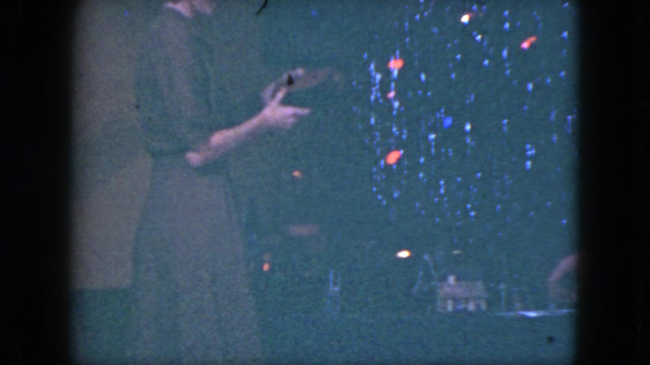 Woman standing beside a miniature train set in a dim room with colored holiday lights.