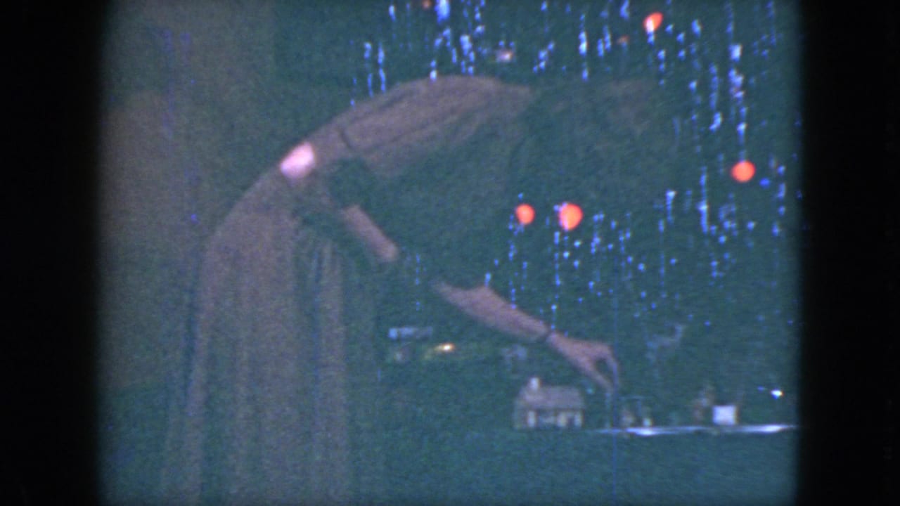 Woman in a long dress bends over a toy train table near tinsel and colored lights.