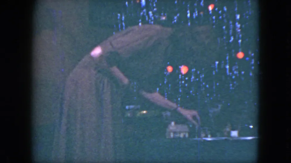 In 1951 Cleveland home-movie footage, a woman in a long dress leans over a miniature train layout beside tinsel and Christmas lights.