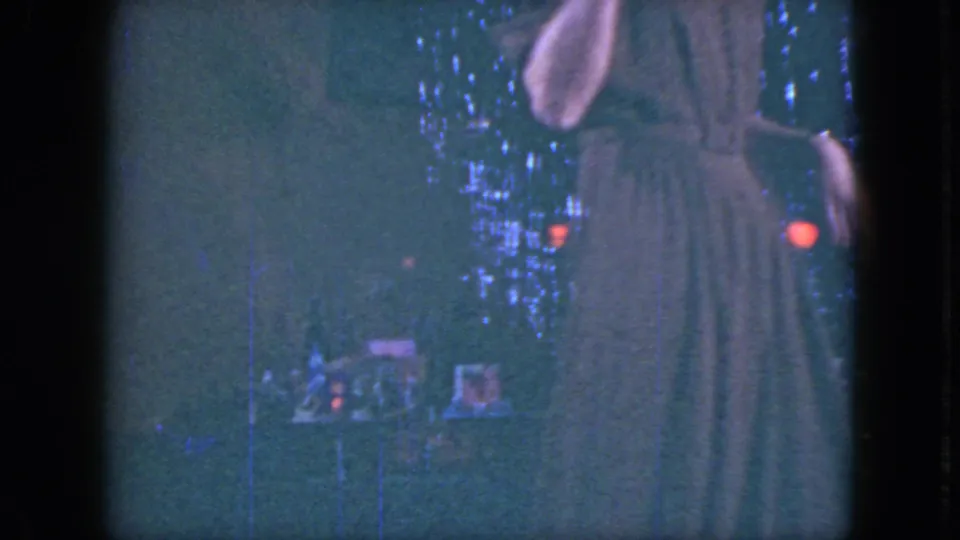A man in a dark suit appears beside the model train display, with tinsel and colored holiday lights in the background.