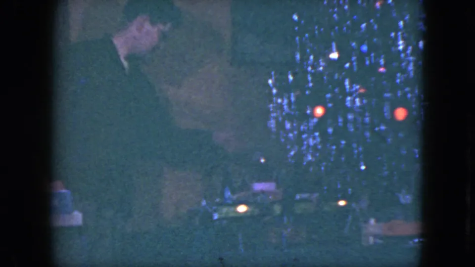 The man gestures near the toy train layout in the 1951 Cleveland holiday scene, partly obscured by low light and film grain.