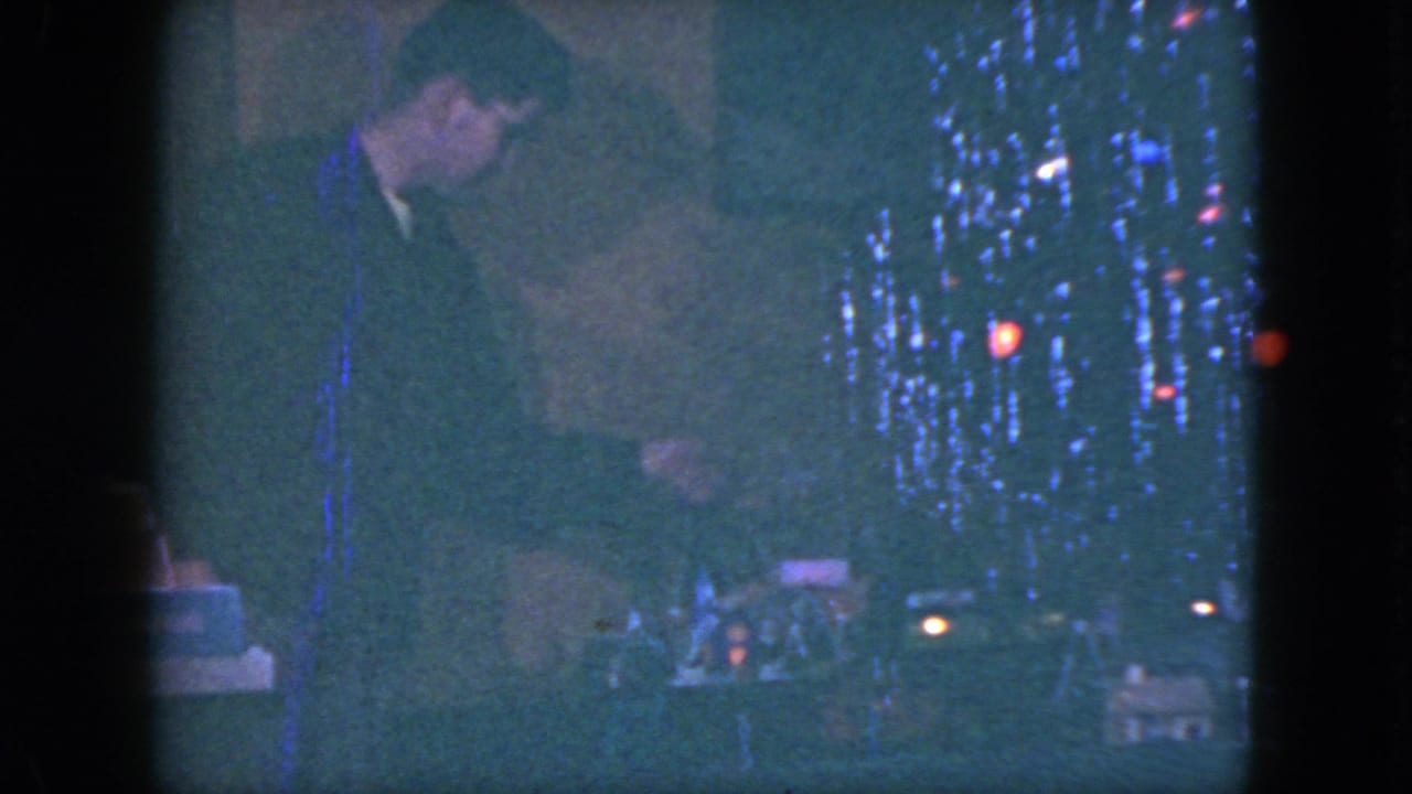 Suited man bends beside a lit Christmas tree and small train-set display in a dim room.