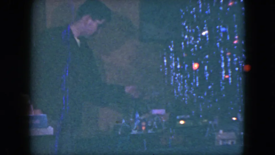 In a 1951 Cleveland home-movie frame, a suited man leans near a decorated Christmas tree and miniature train-set display.