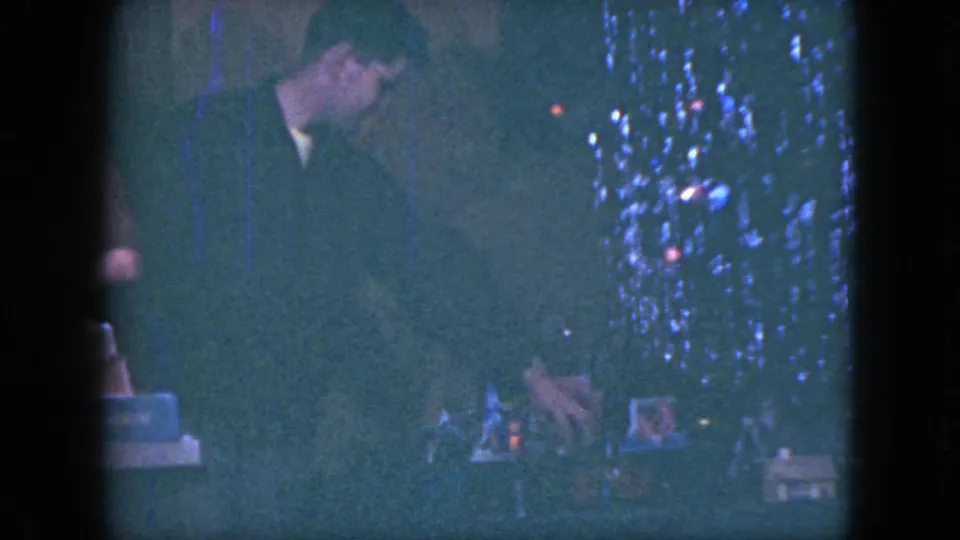 The suited man bends close to the Christmas train set, with tree decorations and small display pieces visible in the 1951 Cleveland footage.