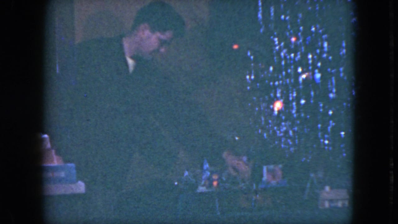 In a 1951 Cleveland home-movie frame, a man stands near a decorated Christmas tree while looking down toward a toy train set.