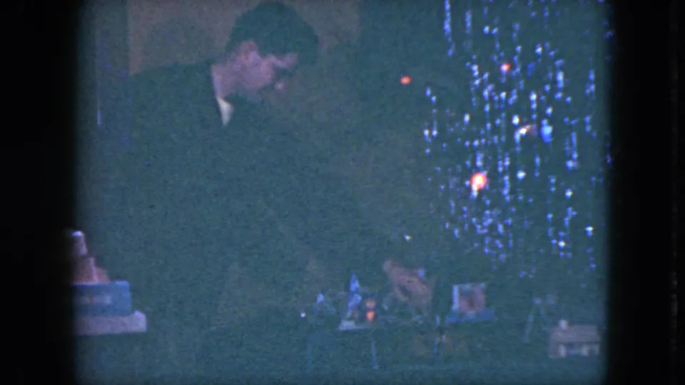 In a 1951 Cleveland home-movie frame, a man stands near a decorated Christmas tree while looking down toward a toy train set.