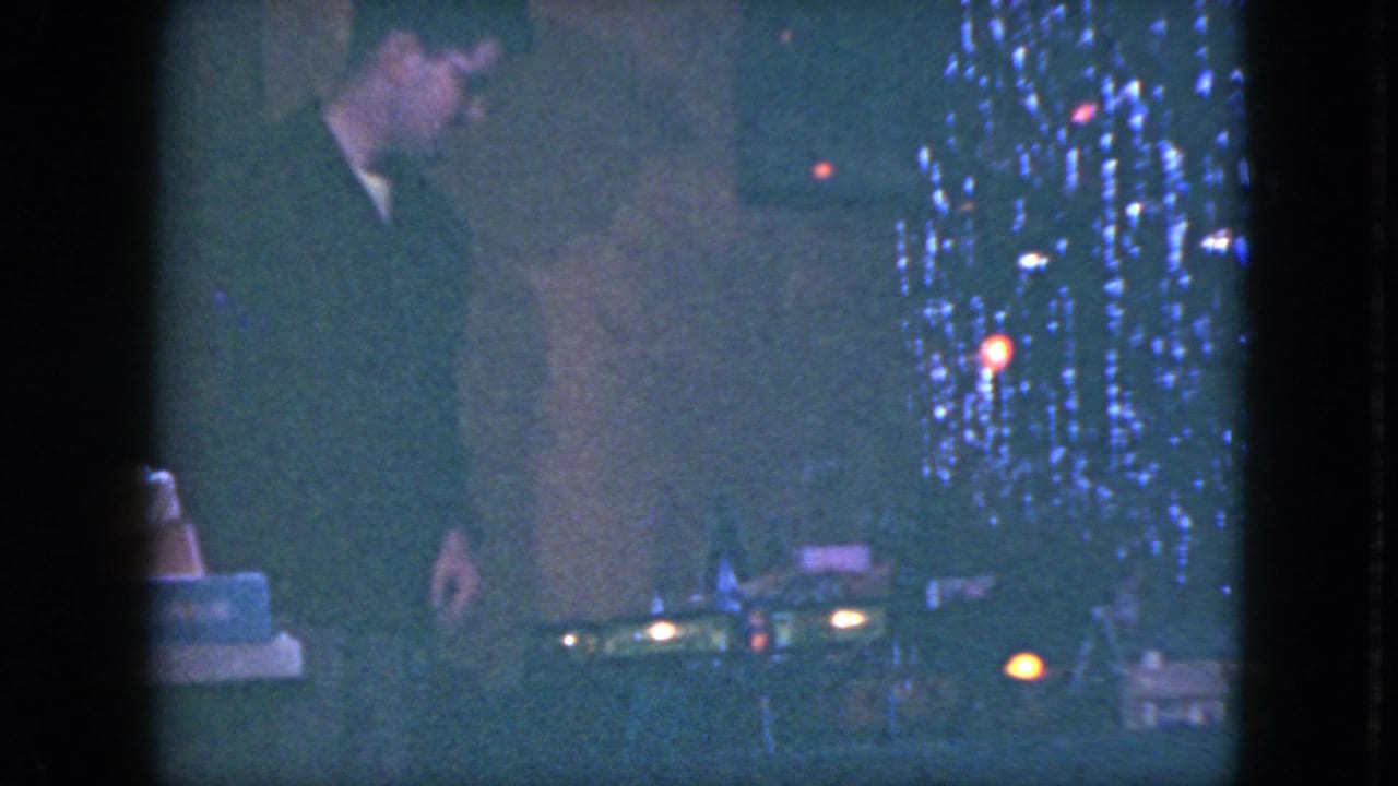 A man in a dark sweater looks toward the table-top train set beside a lit Christmas tree in 1951 Cleveland holiday footage.