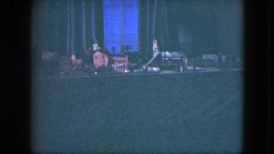 1951 Cleveland home-movie frame of a miniature railroad scene with tiny structures, track, and a blue-lit backdrop in low light.
