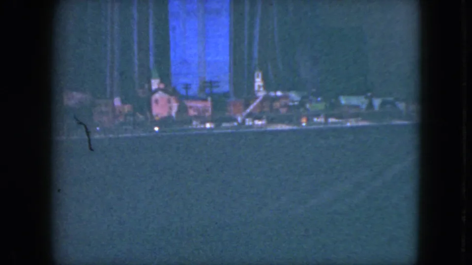 1951 Cleveland home-movie frame of a model railroad scene with small structures, track, and a blue-lit backdrop under dim interior lighting.