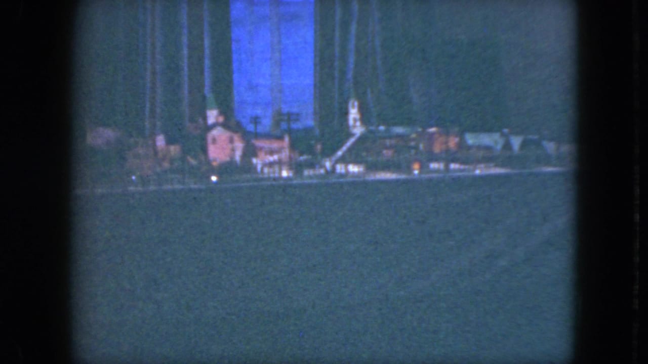 1951 Cleveland 8mm frame showing a miniature train-layout village with small buildings, bare model trees, tiny lights, and a dark backdrop.
