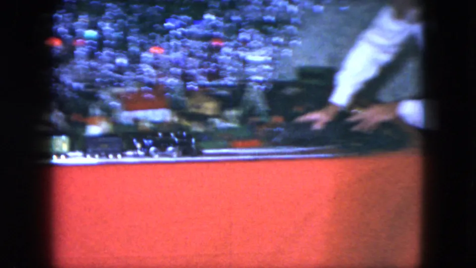 A partially visible adult reaches toward the model train setup beneath the Christmas tree in this 1951 Cleveland home movie frame.