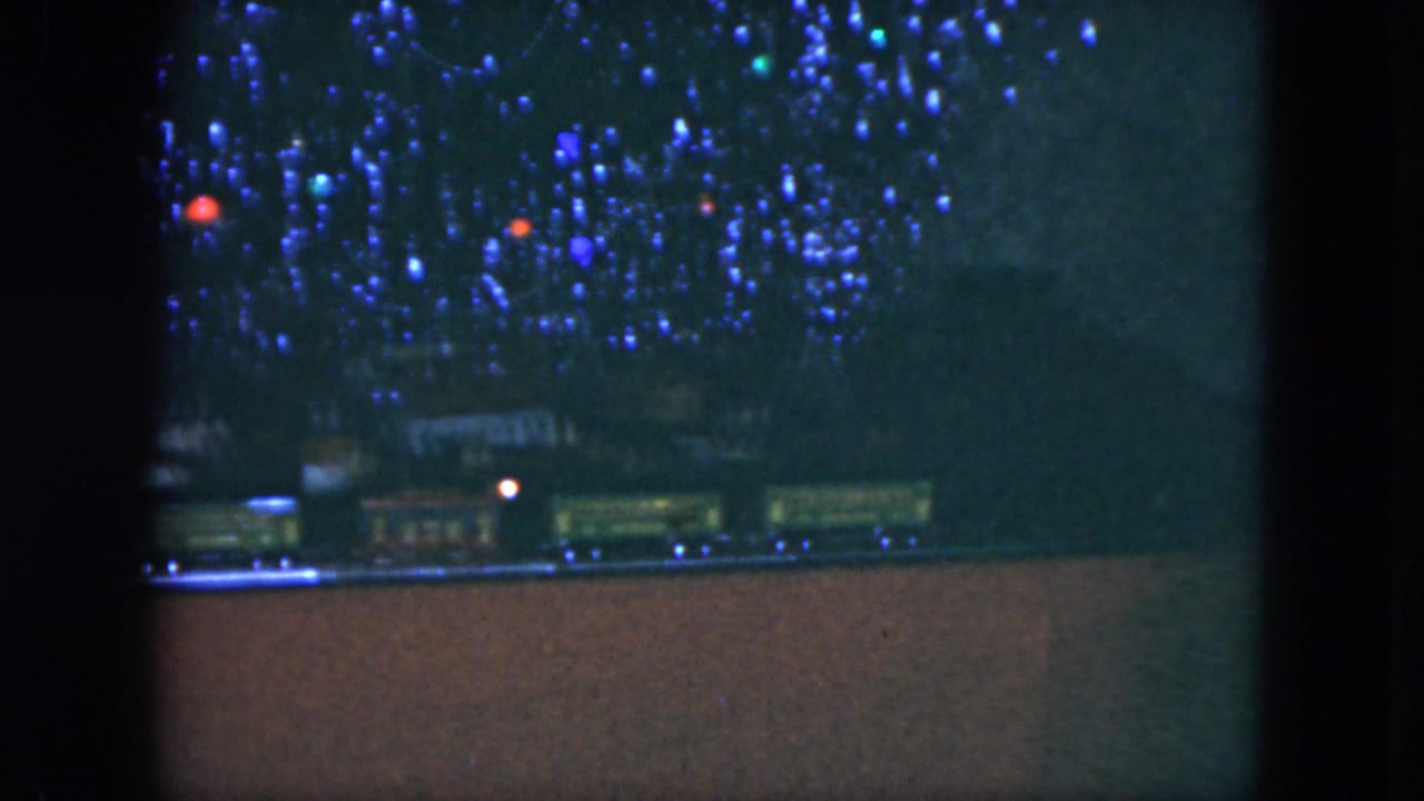 1951 Cleveland home-movie frame showing blue holiday lights over a small train display, viewed through dark interior framing.