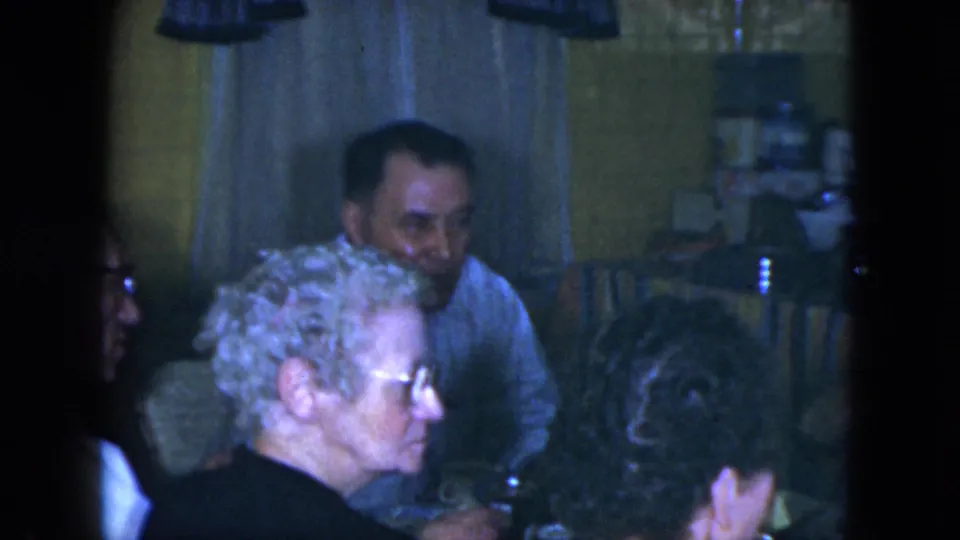 1951 Cleveland home-movie frame showing adults seated around a kitchen table, with cups, curtains, and yellow wall tile visible.