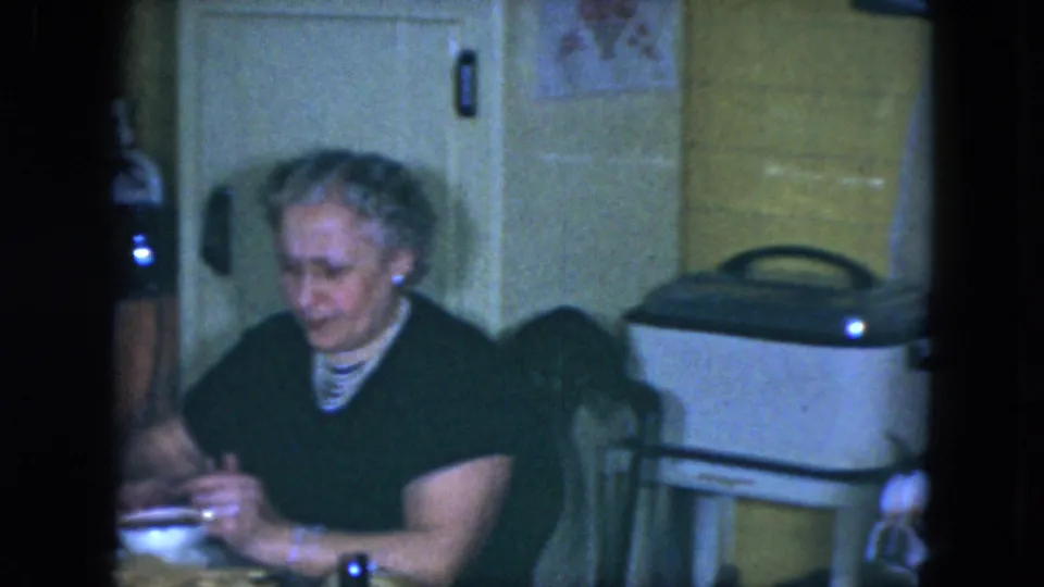 1951 Cleveland home-movie frame showing an older woman at the table beside yellow tile and a lidded kitchen appliance.