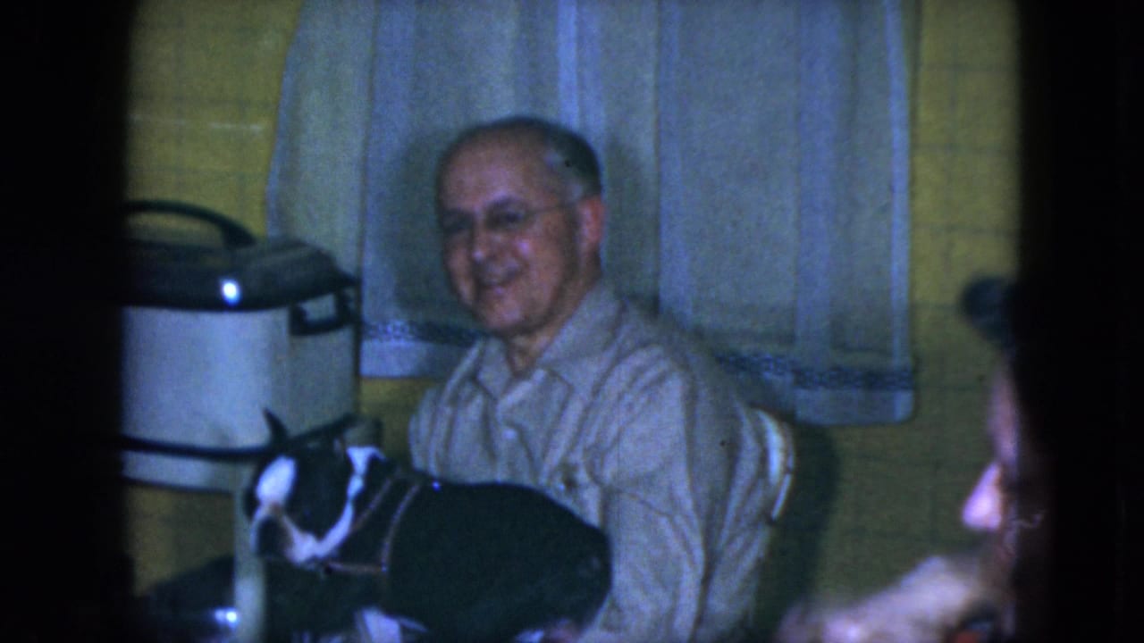 Older man with glasses sits in a kitchen with a black-and-white dog on his lap.