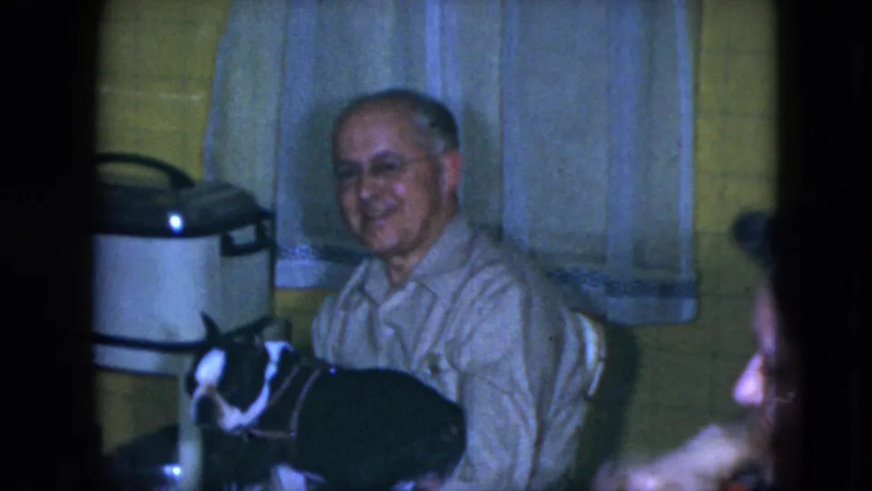 In a 1951 Cleveland home-movie frame, an older man with glasses smiles while seated beside a black-and-white dog in a yellow tiled kitchen.